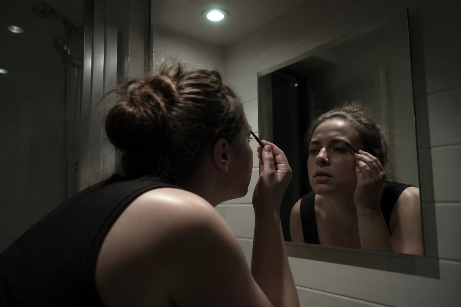 Applying makeup under a single harsh bathroom downlight, face half in shadow